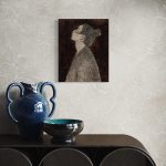 Kulmaleri af kvinde i profil. Kvinden har en høj knold og kigger op mod himlen med lukkede øjne. Hun bærer en oversize sort jakke og baggrunden er mørkebrun. Giclee tryk indrammet med bred sort passepartout hængende i en spisestue.